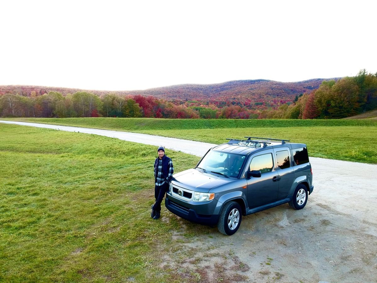 Manny leaning on a grey Honda Element in a field below hills in autumn colours